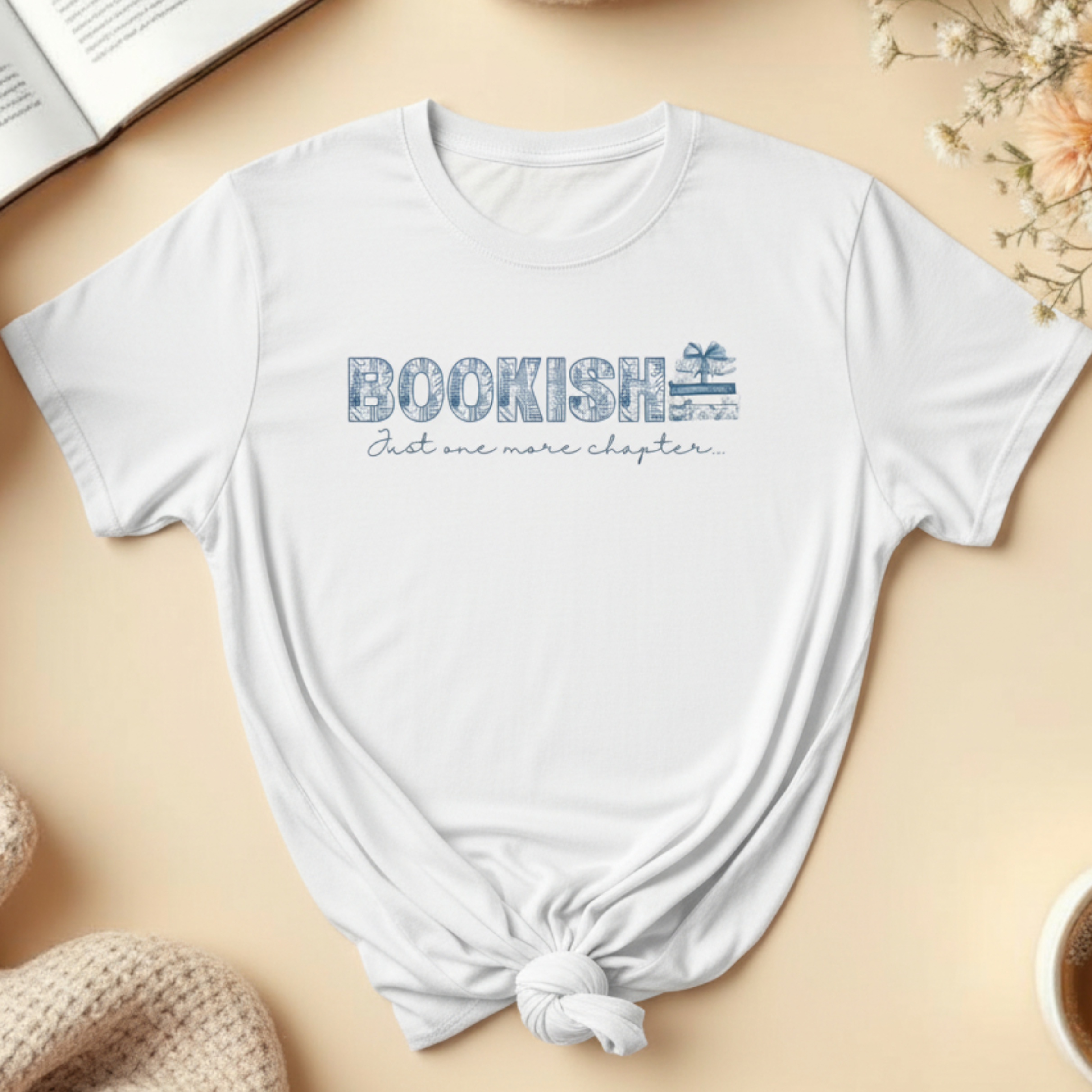 White t-shirt with 'BOOKISH Just one more chapter' text on a beige surface with books, flowers, and a cup.
